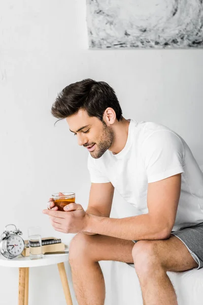 Happy handsome man in pajamas holding cup of tea in bedroom — Stock Photo