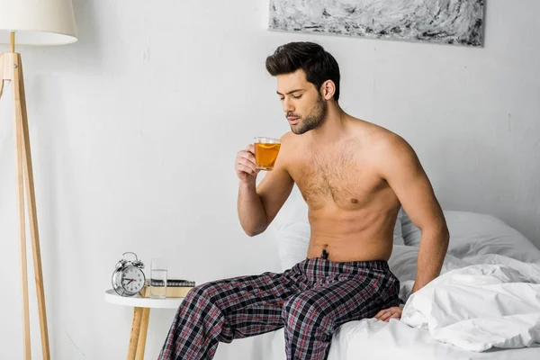 Shirtless man holding cup of tea and sitting on bed — Stock Photo