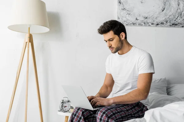 Bello giovane utilizzando il computer portatile mentre seduto in camera da letto — Foto stock