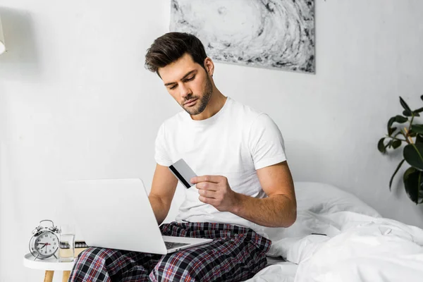 Hombre guapo de compras en línea con tarjeta de crédito y portátil mientras está sentado en el dormitorio - foto de stock