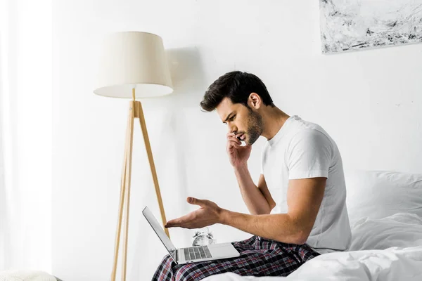 Тривожний чоловік розмовляє на смартфоні і використовує ноутбук на ліжку — стокове фото