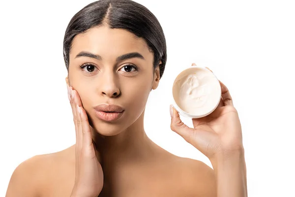 Attraente giovane donna afro-americana in possesso di contenitore con crema e toccare il viso isolato su bianco — Foto stock