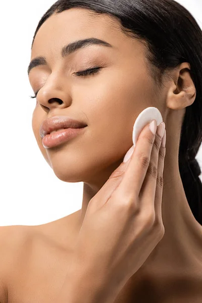 Vista da vicino di bella ragazza afro-americana con gli occhi chiusi pulizia viso con disco di cotone isolato su bianco — Foto stock