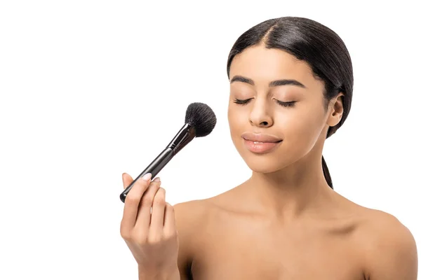 Belle femme afro-américaine souriante aux yeux fermés tenant une brosse cosmétique et appliquant un maquillage isolé sur blanc — Photo de stock