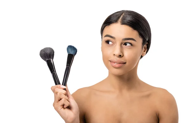 Bela mulher afro-americana sorridente segurando pincéis de maquiagem e olhando para longe isolado no branco — Fotografia de Stock