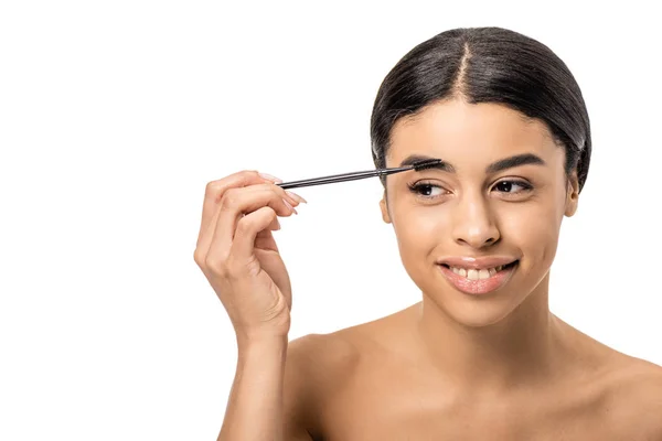 Bela menina americana africana sorridente pentear sobrancelha com escova e olhando para longe isolado no branco — Fotografia de Stock