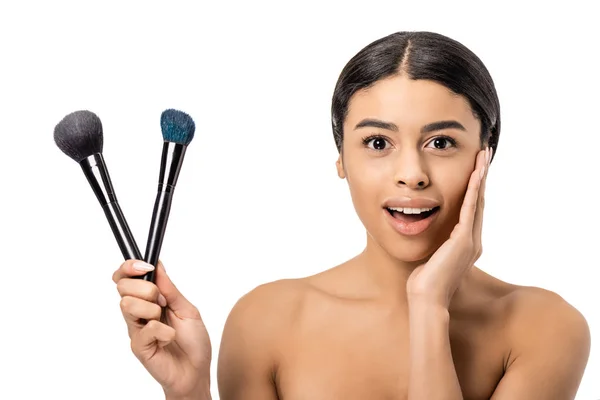 Excited naked african american woman holding makeup brushes and smiling at camera isolated on white — Stock Photo