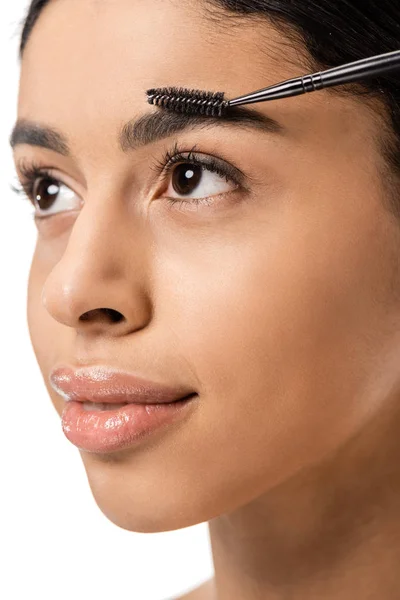 Atractiva chica afroamericana sonriente peinando cejas con cepillo y mirando hacia otro lado aislado en blanco - foto de stock