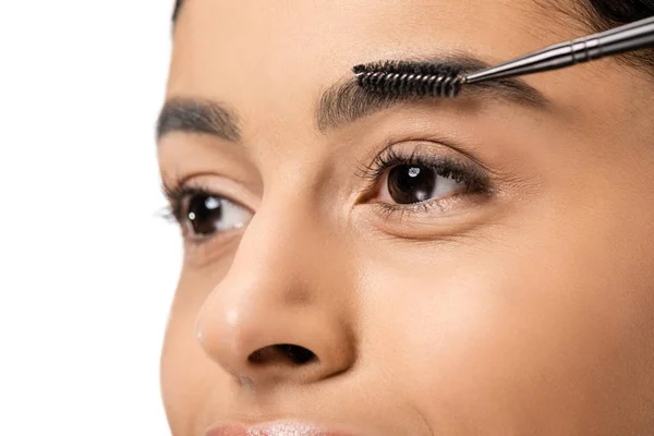 Vista de cerca de la hermosa mujer afroamericana peinando cejas con cepillo y mirando hacia otro lado aislado en blanco - foto de stock