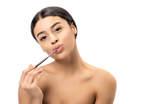 Hermosa mujer afroamericana desnuda aplicando lápiz labial con cepillo cosmético y mirando a la cámara aislada en blanco - foto de stock