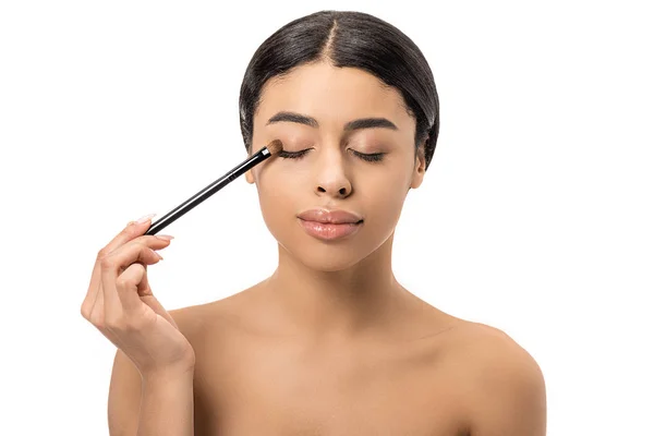 Приваблива брюнетка афроамериканська дівчина з закритими очима наносить тіні з косметичним пензлем ізольовано на білому — стокове фото
