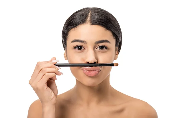 Atractiva mujer afroamericana joven sosteniendo cepillo de maquillaje en los labios y mirando a la cámara aislada en blanco - foto de stock