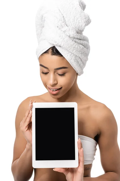 Приваблива усміхнена афроамериканська дівчина з рушником на голові дивиться вниз на цифровий планшет з порожнім екраном ізольовано на білому — стокове фото