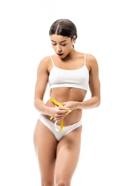 Chocado jovem afro-americano mulher em roupa interior medir cintura com fita e olhando para baixo isolado no branco — Fotografia de Stock