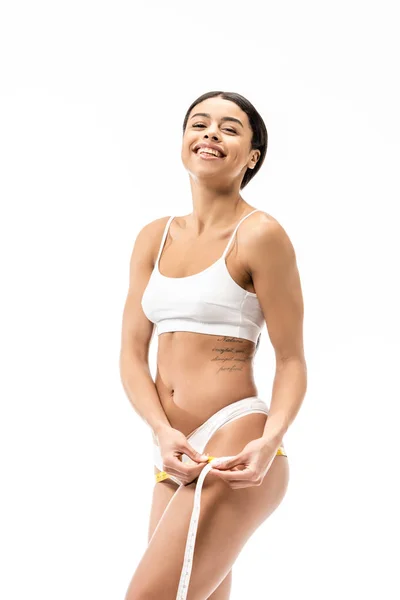 Attractive african american girl in underwear measuring hips with tape and smiling at camera isolated on white — Stock Photo