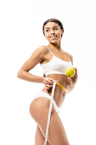 Hermosa mujer afroamericana feliz en ropa interior sosteniendo manzana y cinta métrica aislada en blanco - foto de stock