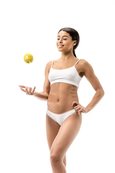 Beautiful smiling african american girl in underwear standing with hand on hip and throwing green apple isolated on white — Stock Photo