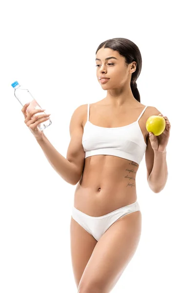 Mince jeune afro-américaine en sous-vêtements tenant bouteille d'eau et pomme verte isolé sur blanc — Photo de stock