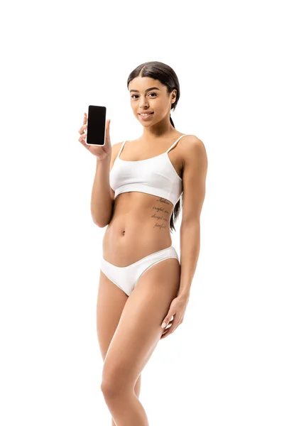 Hermosa chica afroamericana en ropa interior sosteniendo teléfono inteligente con pantalla en blanco y sonriendo a la cámara aislada en blanco - foto de stock