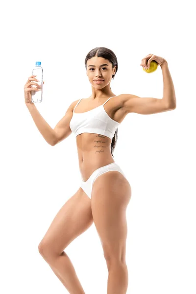 Sportive afro-américaine fille en sous-vêtements tenant bouteille d'eau avec pomme verte et montrant les muscles isolés sur blanc — Photo de stock