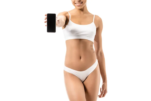 Обрезанный снимок улыбающейся африканской девушки в белых трусах, держащей смартфон с пустым экраном, изолированным на белом — стоковое фото