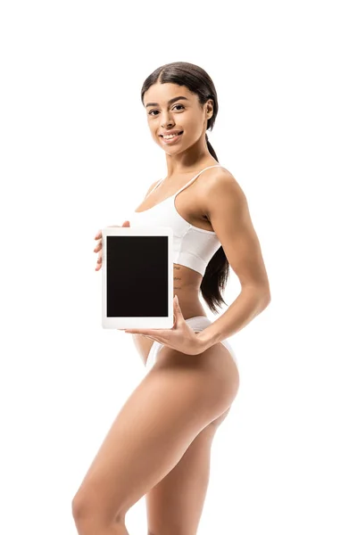 Красивая африканская американка в белом нижнем белье держит цифровой планшет с черным экраном и улыбается на камеру изолированы на белом — стоковое фото