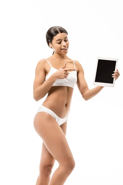 Hermosa sonriente joven afroamericana mujer en ropa interior apuntando con el dedo a la tableta digital aislado en blanco - foto de stock