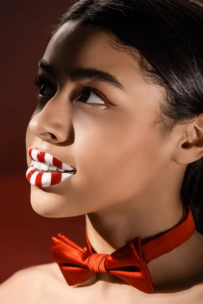 Schöne junge afrikanisch-amerikanische Frau mit roter Fliege und kreativem Make-up, die isoliert auf Burgund blickt — Stockfoto
