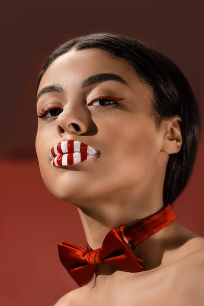 Attractive african american girl with red bow tie and creative makeup looking at camera isolated on burgundy — Stock Photo