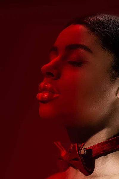 Toned image on young seductive african american woman with closed eyes and red bow tie isolated on burgundy — Stock Photo