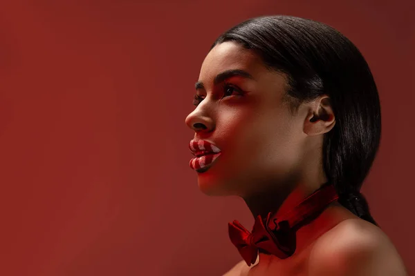 Seitenansicht der schönen nackten afrikanisch-amerikanischen Mädchen mit Fliege und gestreiften Lippen wegschauen isoliert auf Burgund — Stockfoto