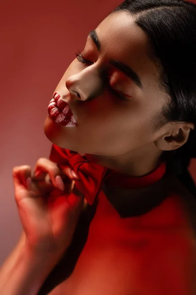 High angle view of beautiful sensual young african american woman with bow tie and striped lips posing with closed eyes isolated on burgundy — Stock Photo