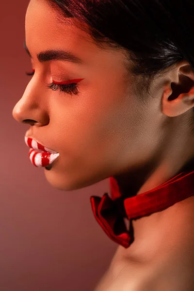 Vue latérale de belle fille africaine américaine sensuelle avec nœud papillon rouge et lèvres rayées isolées sur bordeaux — Photo de stock