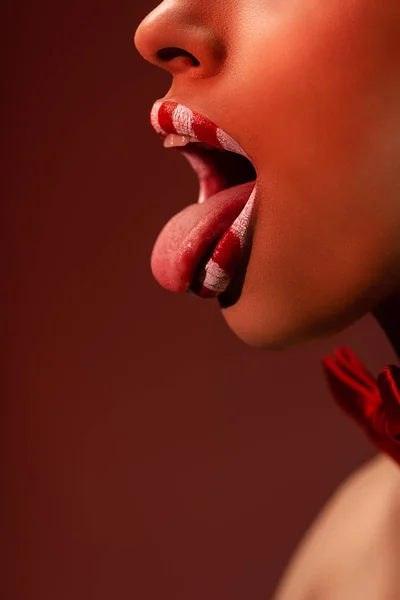 Plan recadré de séduisante femme afro-américaine aux lèvres rouges et blanches montrant la langue isolée sur bordeaux — Photo de stock