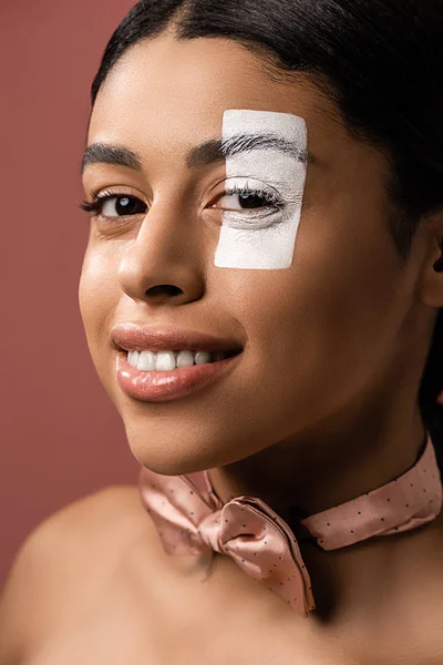 Bela jovem mulher americana africana com gravata borboleta e tinta branca no rosto sorrindo para a câmera isolada em marrom — Fotografia de Stock