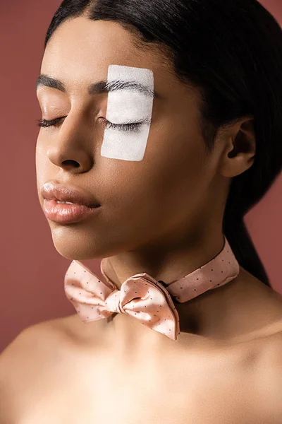 Hermosa joven afroamericana mujer con pajarita y pintura blanca en la cara posando con los ojos cerrados aislados en marrón - foto de stock