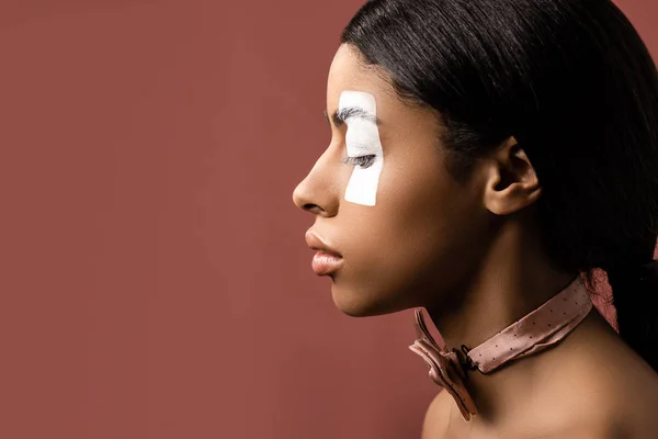 Vue latérale de la femme afro-américaine avec trait de peinture blanche sur les yeux et noeud papillon regardant vers le bas isolé sur brun — Photo de stock