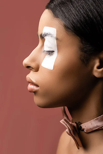Vista lateral de hermosa mujer afroamericana con trazo de pintura blanca en el ojo y pajarita aislada en marrón - foto de stock
