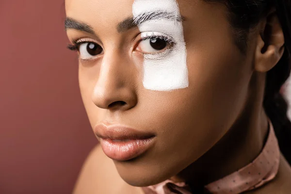 Belle femme afro-américaine avec trait de peinture blanche sur les yeux en regardant la caméra isolée sur brun — Photo de stock