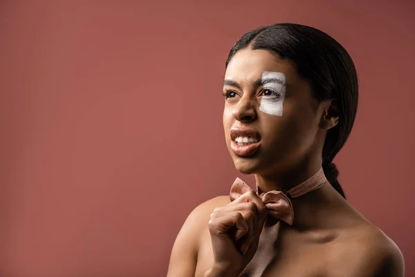 Angry naked african american woman with white paint stroke on eye holding bow tie and looking away isolated on brown — Stock Photo