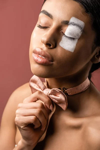 Sensual african american woman with closed eyes and white paint on face adjusting bow tie isolated on brown — Stock Photo