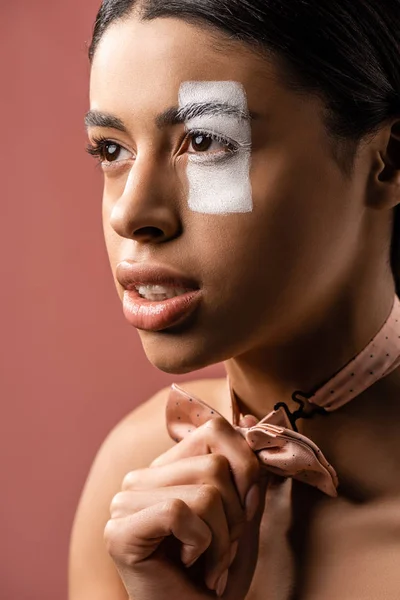 Belle femme afro-américaine focalisée avec noeud papillon et peinture blanche sur le visage regardant loin isolé sur brun — Photo de stock