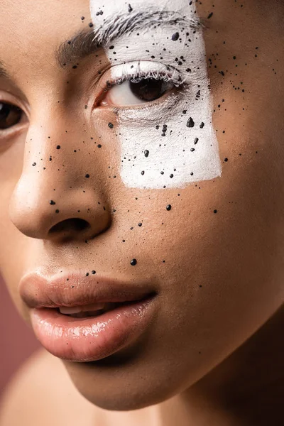 Vue rapprochée de femme sensuelle afro-américaine avec art corporel créatif sur le visage en regardant la caméra isolée sur brun — Photo de stock