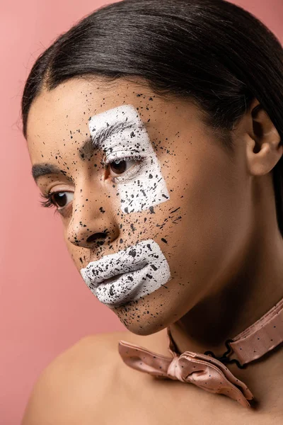 Attraktive nachdenkliche afrikanisch-amerikanische Mädchen mit Fliege und Farbe im Gesicht sieht weg isoliert auf rosa — Stockfoto