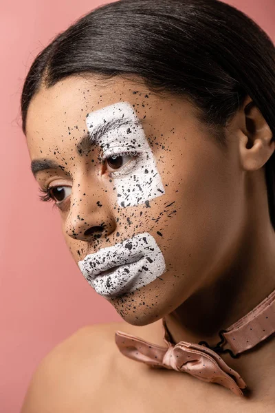 Atractiva chica afroamericana pensativa con pajarita y pintura en la cara mirando hacia otro lado aislado en rosa - foto de stock