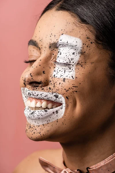 Mujer afroamericana feliz con el arte artístico del cuerpo en la cara riendo con los ojos cerrados aislados en rosa - foto de stock