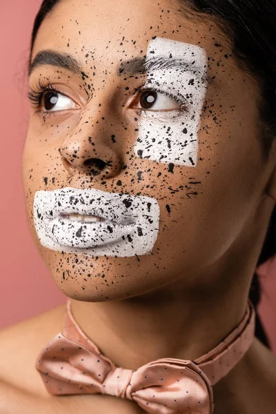 Hermosa mujer afroamericana con pintura blanca y salpicaduras marrones en la cara mirando hacia otro lado aislado en rosa - foto de stock