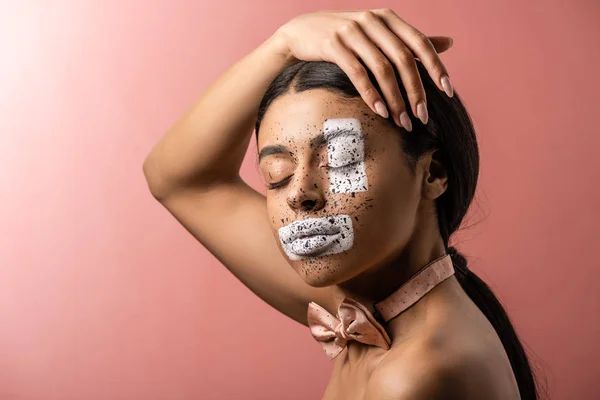 Sensuale donna afroamericana ad occhi chiusi, papillon e vernice sul viso toccando i capelli isolati sul rosa — Foto stock
