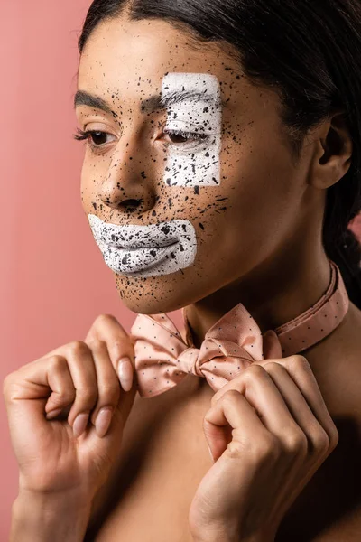 Beautiful african american woman with paint on face adjusting bow tie and looking away isolated on pink — Stock Photo