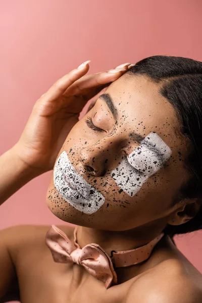 Bela mulher americana africana sensual com tinta no rosto e gravata borboleta tocando cabelo isolado em rosa — Fotografia de Stock